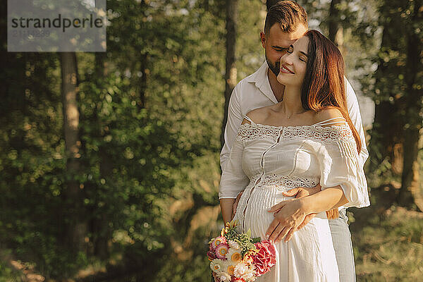 Husband embracing pregnant wife in white dress near meadow