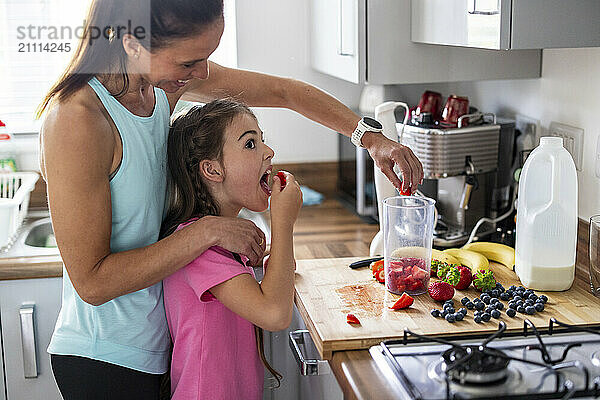 Daughter eating strawberries with mother making milkshake at home