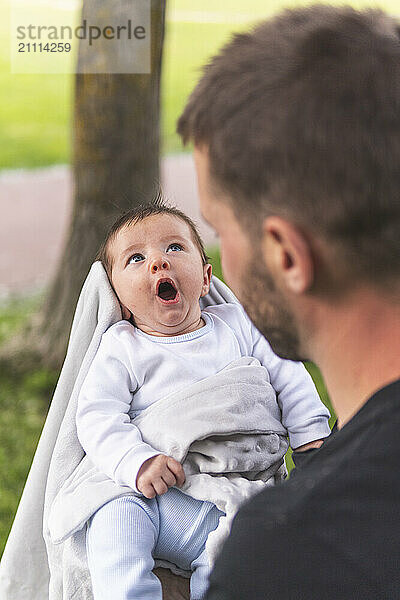 Father looking at baby boy yawning at playground