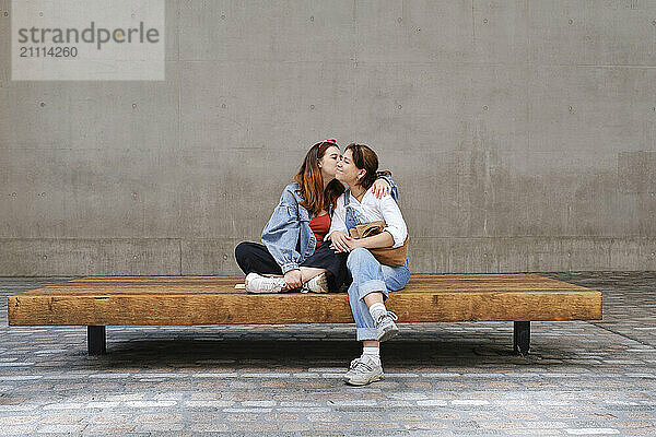 Affectionate woman kissing girlfriend sitting on bench in front of wall