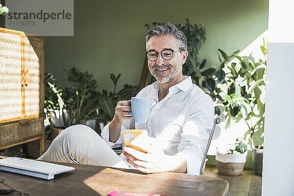 Smiling businessman holding cup and surfing net through smart phone at home office