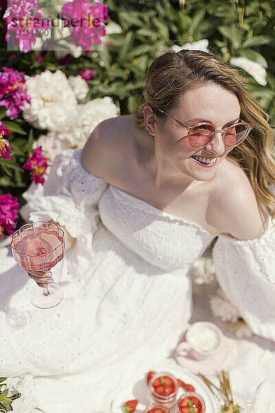 Happy blond woman holding lemonade and sitting near peonies garden