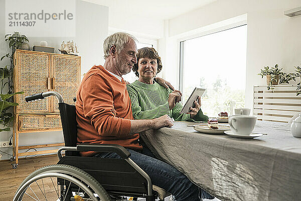 Senior man on wheelchair sitting with wife using digital PC at home