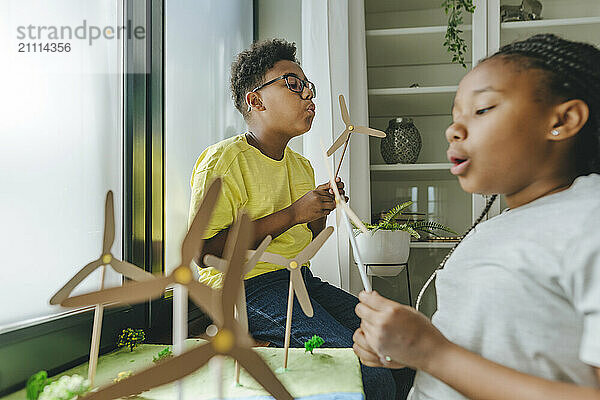 Brother and sister blowing on toy wind turbines at home