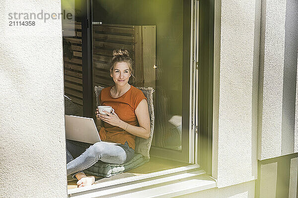 Woman siting by window with laptop and mug