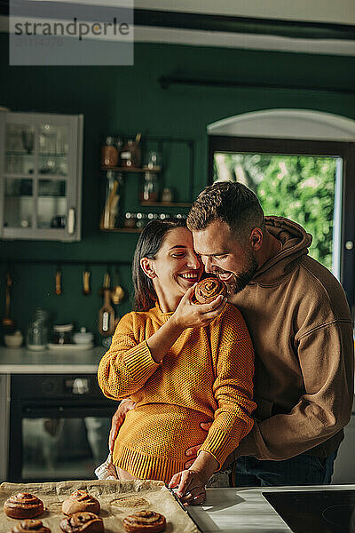 Young pregnant woman feeding cinnamon bun to husband at home