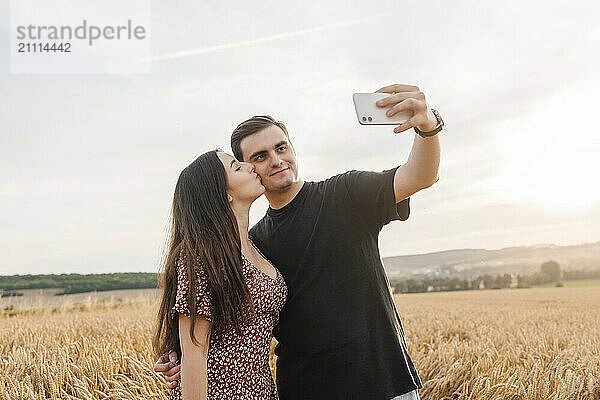 Young woman kissing boyfriend taking selfie through smart phone in field