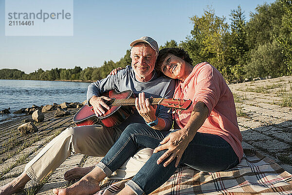 Smiling senior man playing guitar with woman sitting near riverbank on sunny day