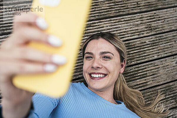 Happy woman taking selfie through smart phone and lying on wooden patio