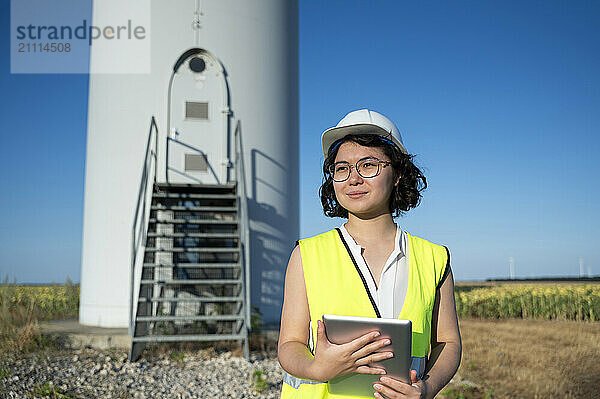 Smiling young maintenance engineer with tablet PC standing at wind farm