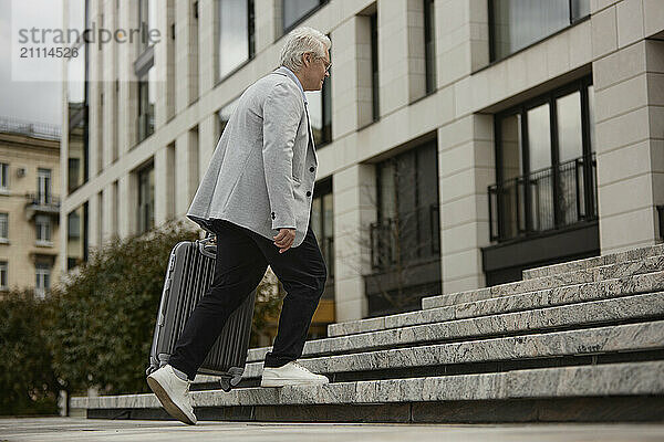 Senior woman with suitcase moving up steps near building in street