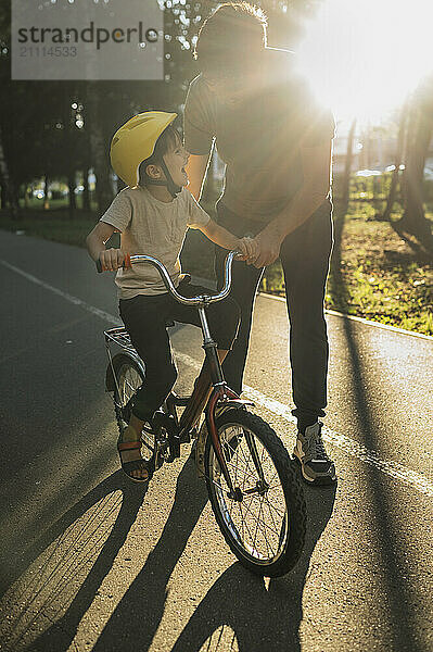 Father helping son in riding bicycle on road during sunny day