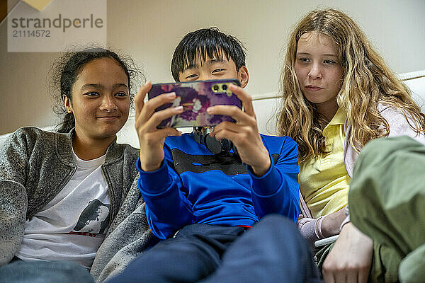 Three children sit closely together  engrossed in a smartphone game  with expressions of concentration and enjoyment.