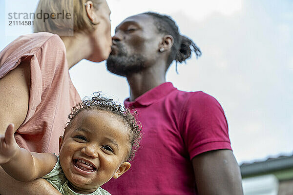 A smiling toddler in a green onesie extends their arm towards the camera as a woman kisses a man on the cheek  both adults in casual clothes with a clear sky in the background.