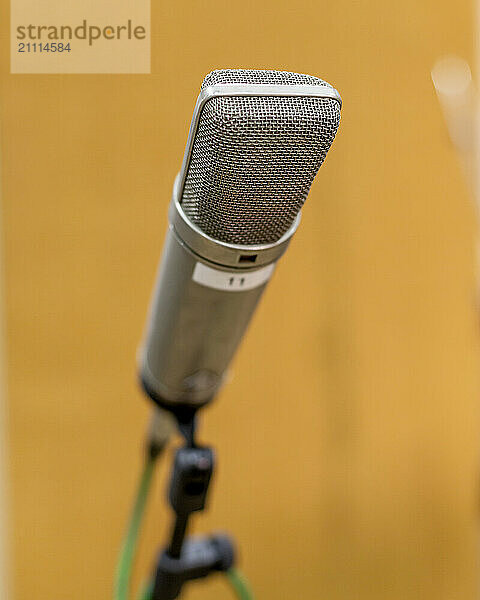 A studio microphone with a silver grille body mounted on a stand  sharp in focus against a blurred orange background.