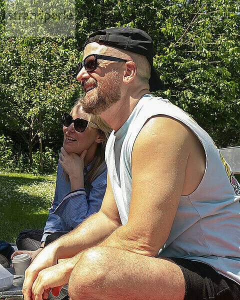 Smiling couple enjoying a sunny day outdoors  the man wearing a tank top and cap seated in front with a woman in sunglasses behind him looking on.