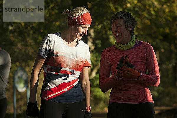 Two women smiling and talking during an outdoor exercise session in a park with warm sunlight.