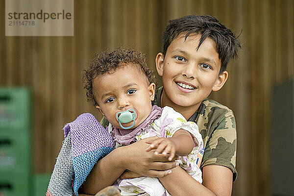 Smiling boy holds a young toddler with a pacifier  both showing a close sibling bond.