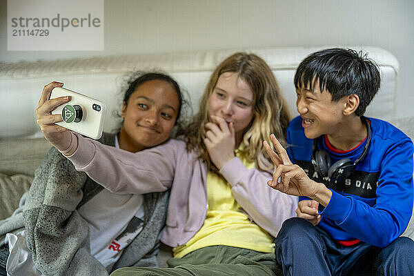 Three kids laughing and taking a selfie on a smartphone while sitting on a couch.