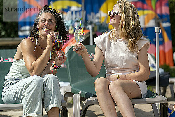 Two women smiling and toasting with cocktails on a sunny beach lounge chair setting.