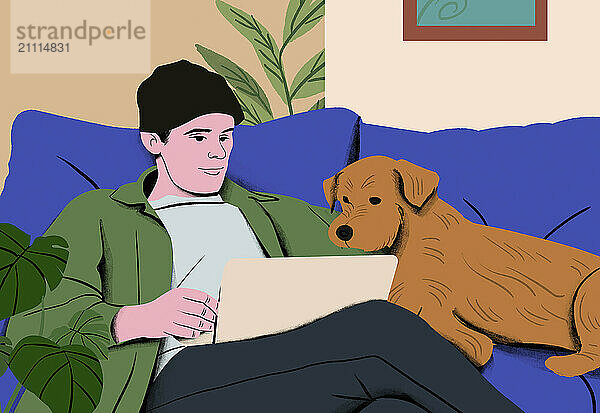 A smiling young man wearing a beanie and green jacket enjoys using his laptop  with a brown dog resting beside him on a blue couch.