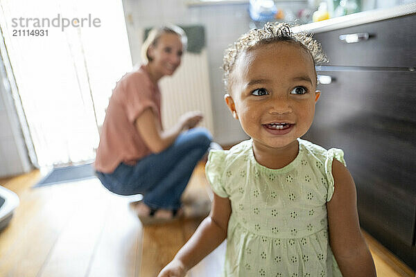 A joyful toddler girl smiles in a sunlit kitchen while a woman kneels in the background.