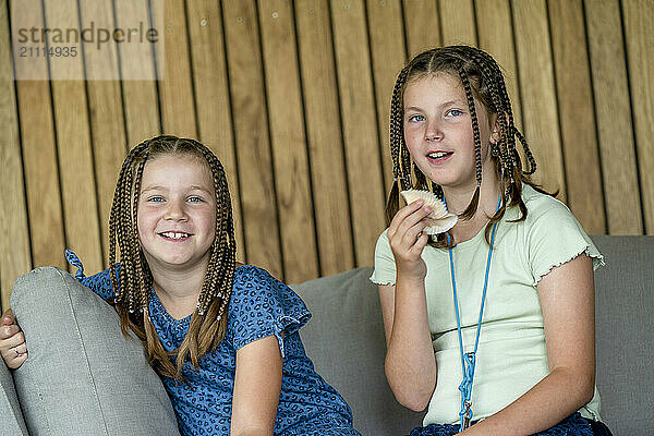 Two girls sitting on a couch  one eating an apple and both smiling with a wooden backdrop.