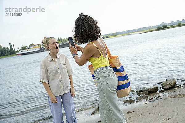 Two women having a joyful conversation by the river with one holding a smartphone and both expressing happiness.