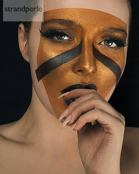 Close-up portrait of a woman with striking brown and black geometric makeup design covering her face and eyelids.