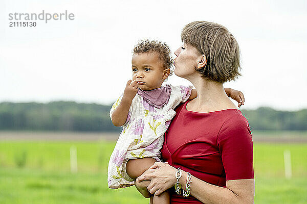 Mother affectionately kissing her baby who looks pensive against a rural backdrop.