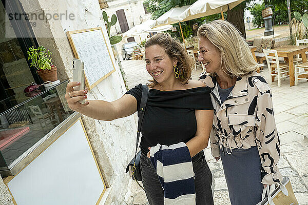 Two women taking a selfie together outside a cafe with umbrellas and greenery in the background.