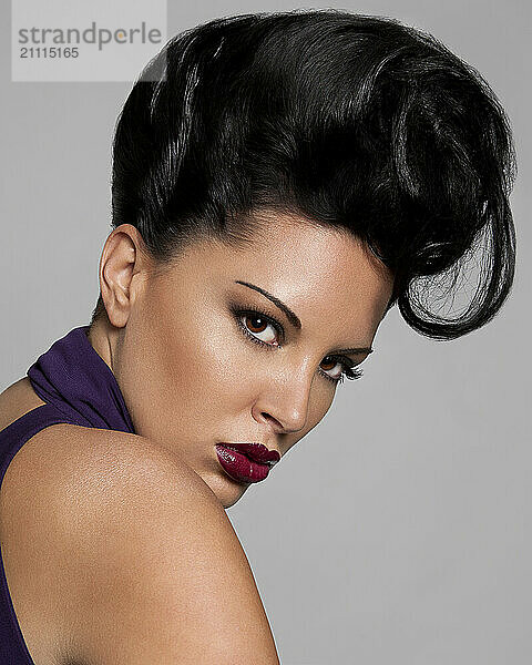 Woman with dramatic makeup and an elaborate hairstyle posing against a gray background.