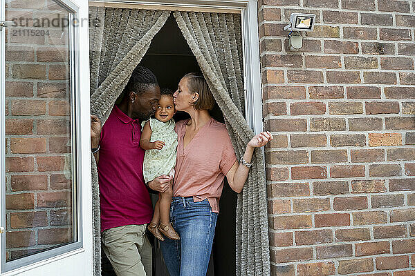 Family with a toddler smiling at each other in a doorway with a brick wall background.