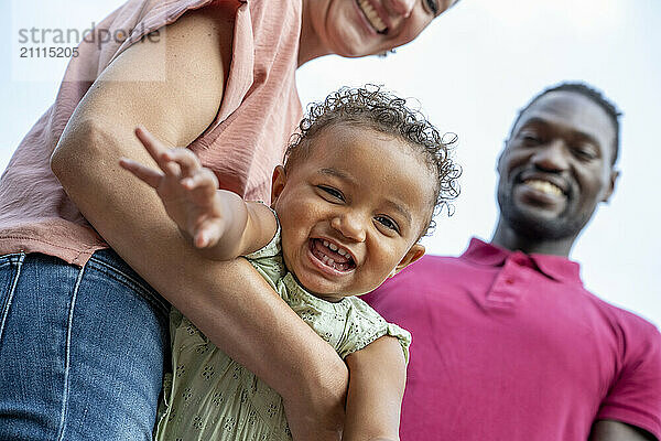 Joyful toddler laughing while being held by smiling woman with a man standing beside them  all enjoying a happy moment together.