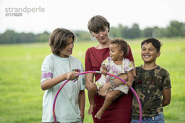 A family enjoys outdoor time together  with a mother holding a toddler  flanked by a smiling boy and another child playing with a hula hoop.