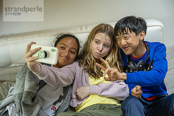 Three kids sitting close together on a couch taking a selfie with a smartphone  smiling and making peace signs.