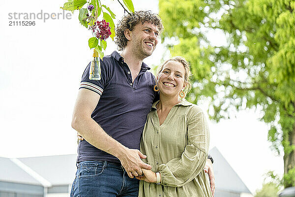 Smiling couple embracing outdoors with a bouquet of flowers.