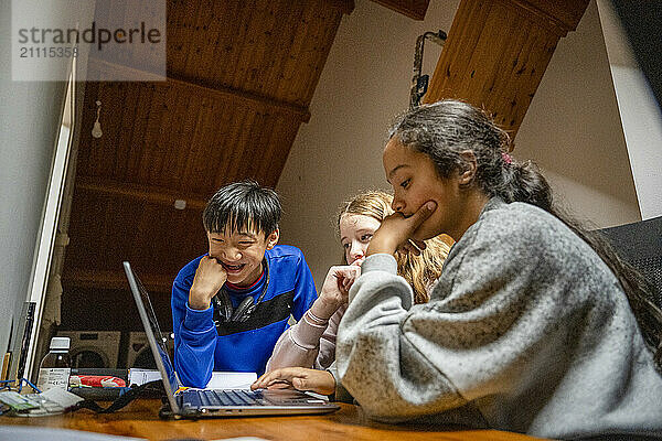 Three children are gathered around a laptop  laughing and enjoying an entertaining moment together in a cozy room with wooden interiors.