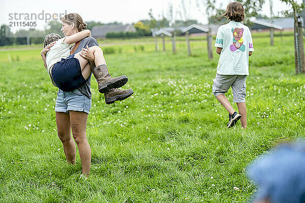 A woman carries a young child on her back while walking through a green field  with another child walking ahead.