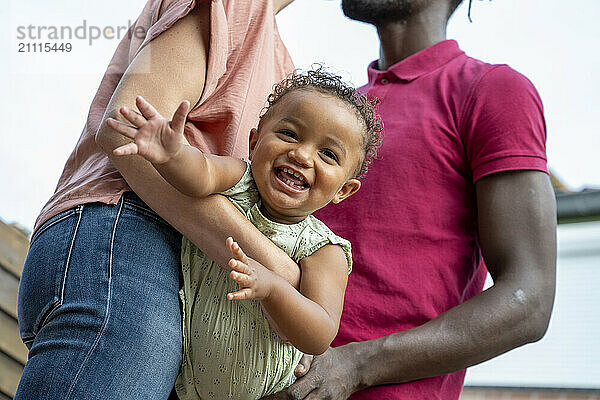 A toddler laughs joyfully while being held by her parents  showcasing a candid family moment outdoors.