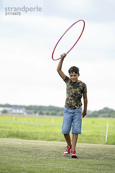 Young boy playing with a pink hula hoop on a grassy field under a cloudy sky.