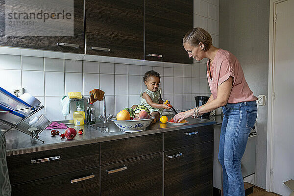 Woman washing dishes at the sink with a young child sitting on the kitchen counter surrounded by fruits and playing with a water tap.