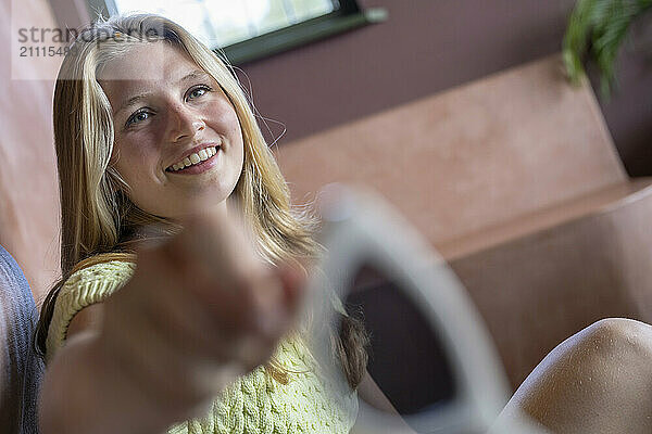 Smiling young woman reaching out towards the camera in a cozy indoor setting.