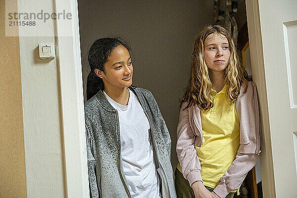 Two teenage girls stand in a doorway  sharing a moment in casual conversation  with one girl smiling subtly.