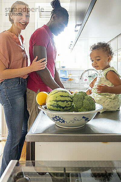 Smiling women standing by a kitchen counter with a toddler  a bowl of fruit in the foreground.