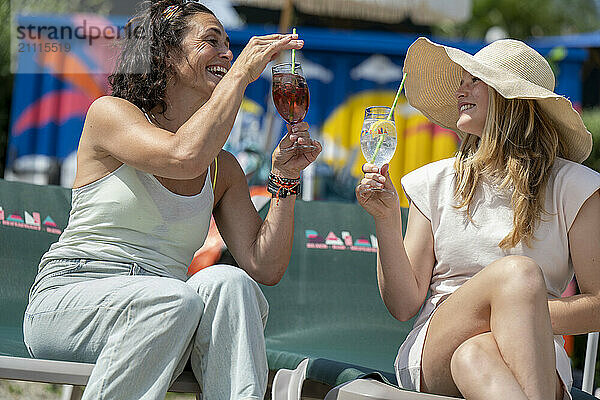 Two women cheer with drinks at a sunny outdoor event  laughing and enjoying each other's company.