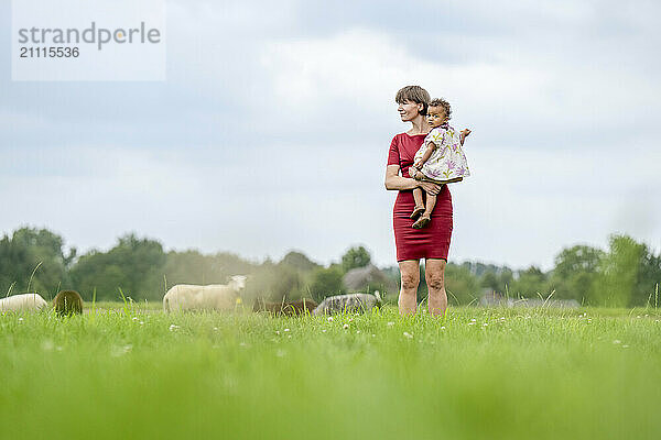 Mother holding her child while standing in a grassy field with sheep in the background.