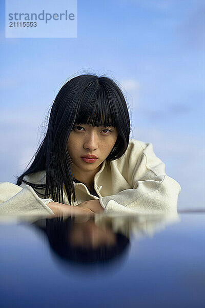 Woman with dark hair lying down  her reflection visible on a glossy surface  against a blue sky backdrop.