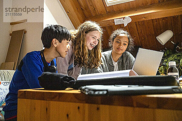 Three teenagers are smiling and looking at a laptop screen together in a warmly lit room with wooden beams.