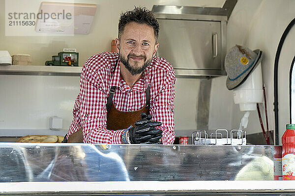 Smiling food truck vendor wearing a red checkered shirt and apron stands at the service window.